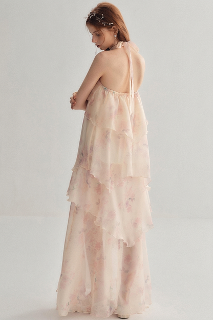 A woman wearing a sheer, pale blush pink halter maxi dress with a soft floral print, shown from the back against a light gray studio background.