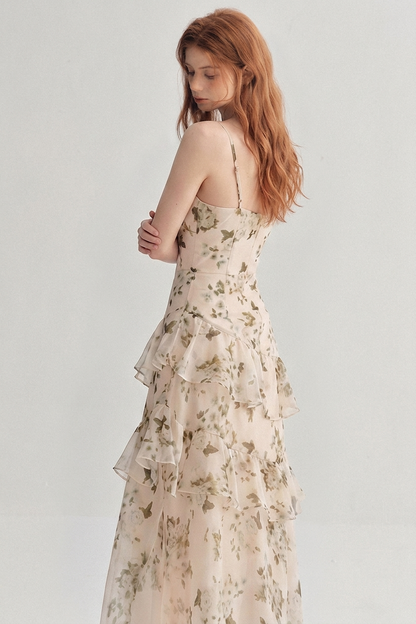 A woman with long wavy auburn hair is shown from the back wearing a sleeveless, tiered maxi dress with a delicate beige and green floral pattern, set against a plain light gray studio background.