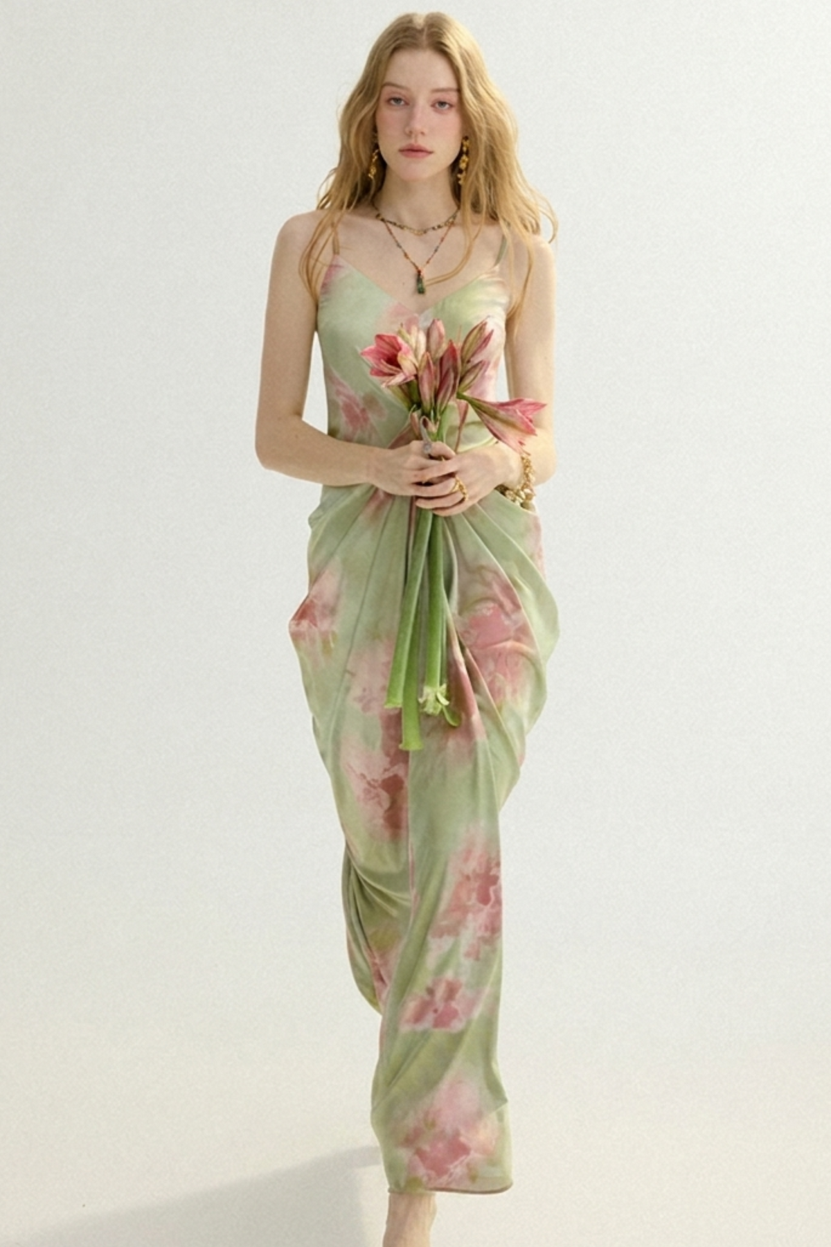 A woman modeling a light sage green maxi dress with a pink floral print, holding a bouquet of pink flowers against a plain white studio background.