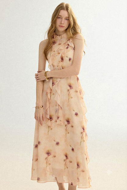 A woman modeling a sleeveless, high-neck midi dress with a ruffled tiered skirt. The dress is a light peach-beige color with a scattered floral print in muted purple and brown tones.