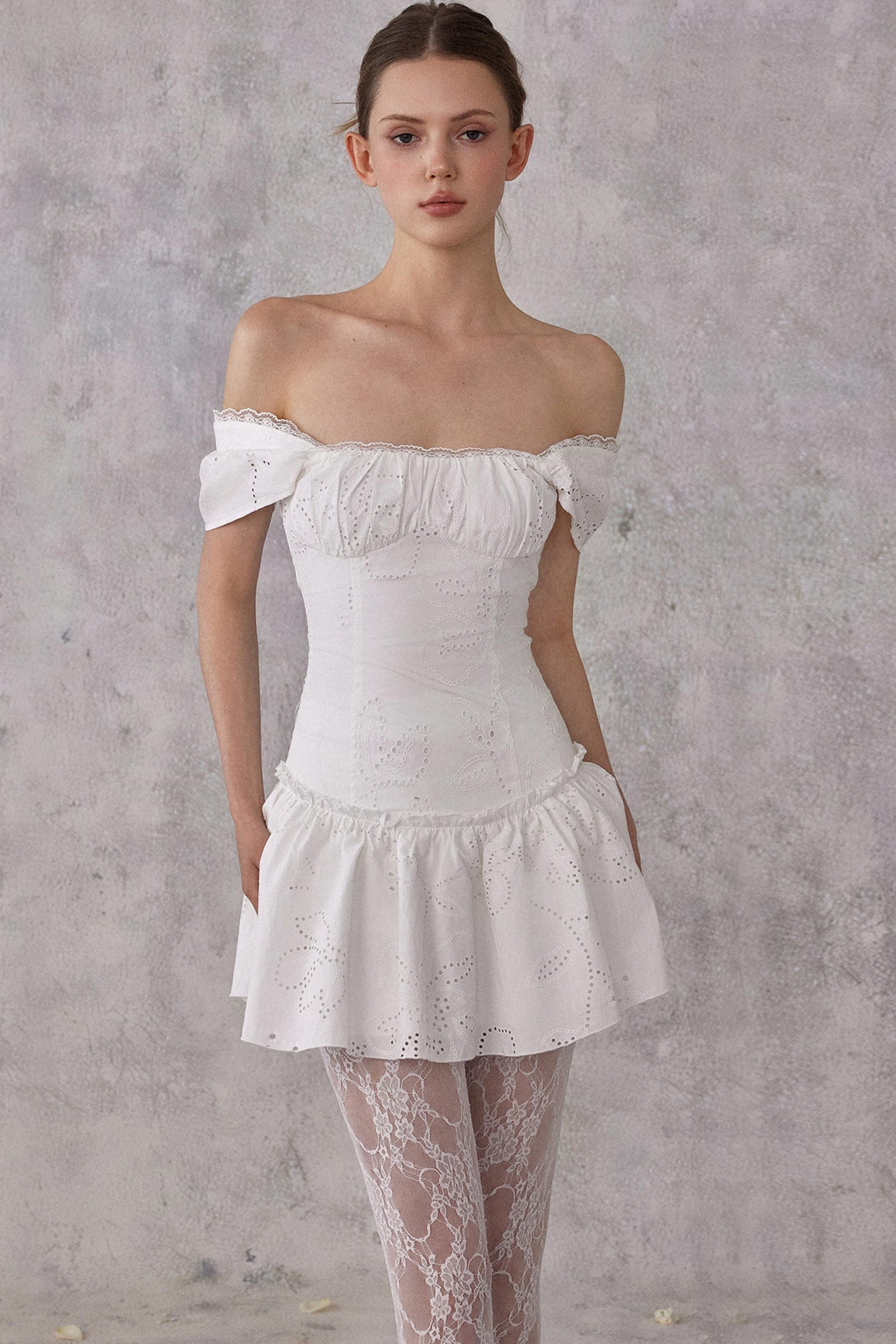 Model styling a romantic white cotton mini dress with eyelet lace details and a smocked pleated waist, worn in a feminine balletcore aesthetic.