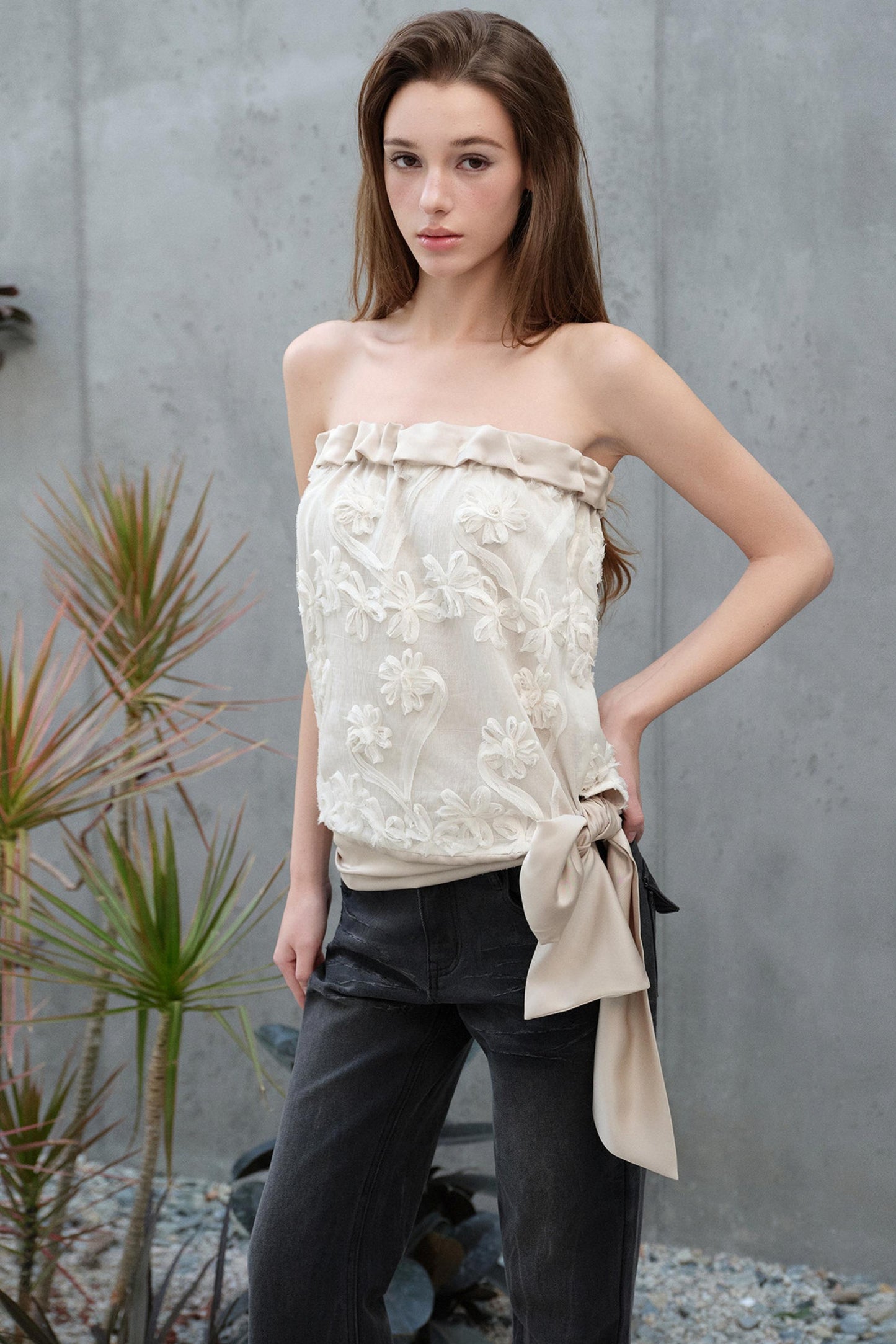 Woman wearing a strapless floral top with a tied front, standing against a gray wall with plants.
