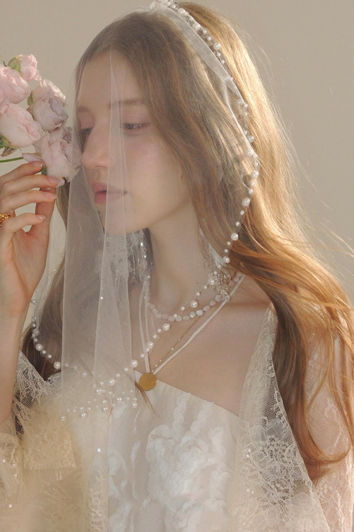 A woman wearing a white lace bridal dress, pearl-beaded headpiece and veil, layered pearl necklaces, and a gold pendant, holding a bouquet of pink roses.