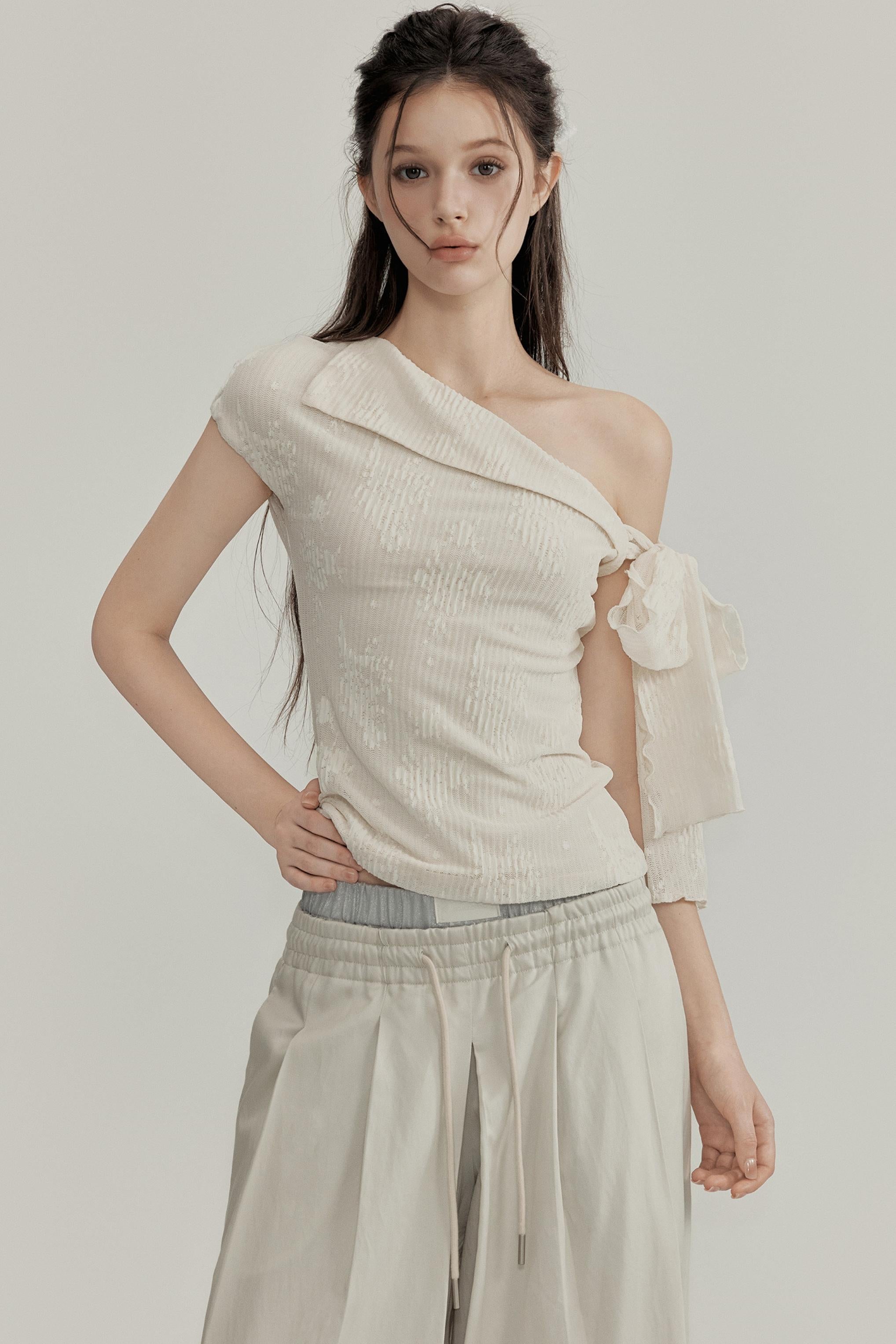 Woman wearing a one-shoulder cream top and beige pants on a plain background