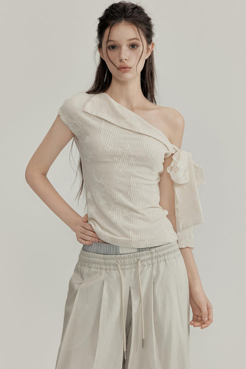 Woman wearing a one-shoulder cream top and beige pants on a plain background