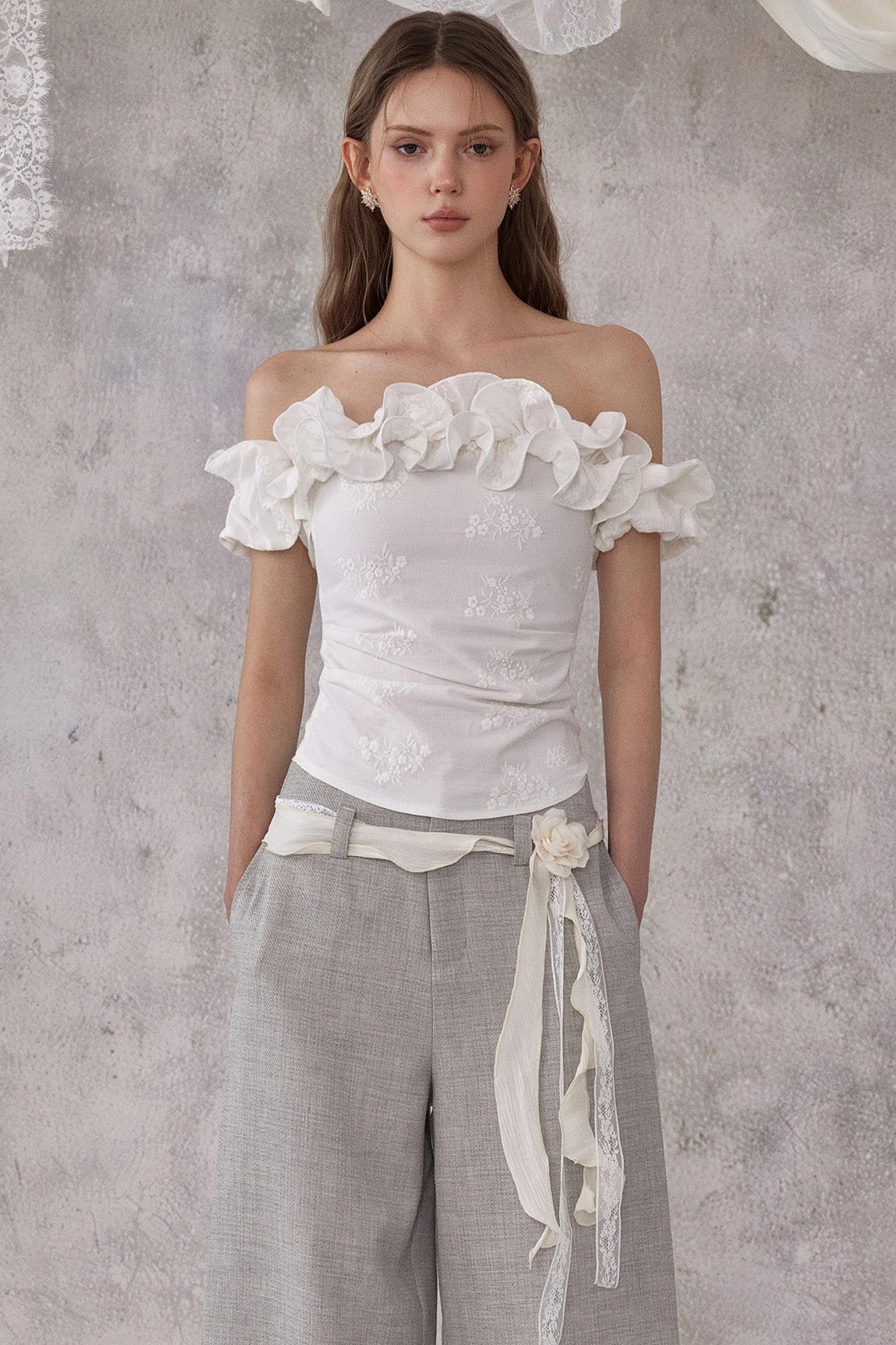 Woman wearing a white off-shoulder top with floral details and gray pants against a textured gray wall.