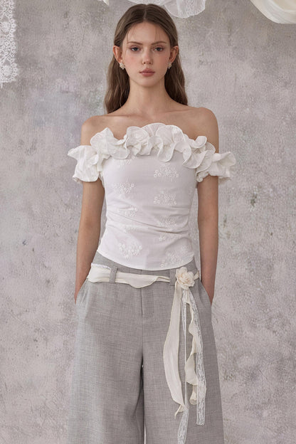 Woman wearing a white off-shoulder top with floral details and gray pants against a textured gray wall.