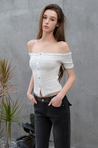 Woman wearing a white off-shoulder top and black pants against a gray wall.