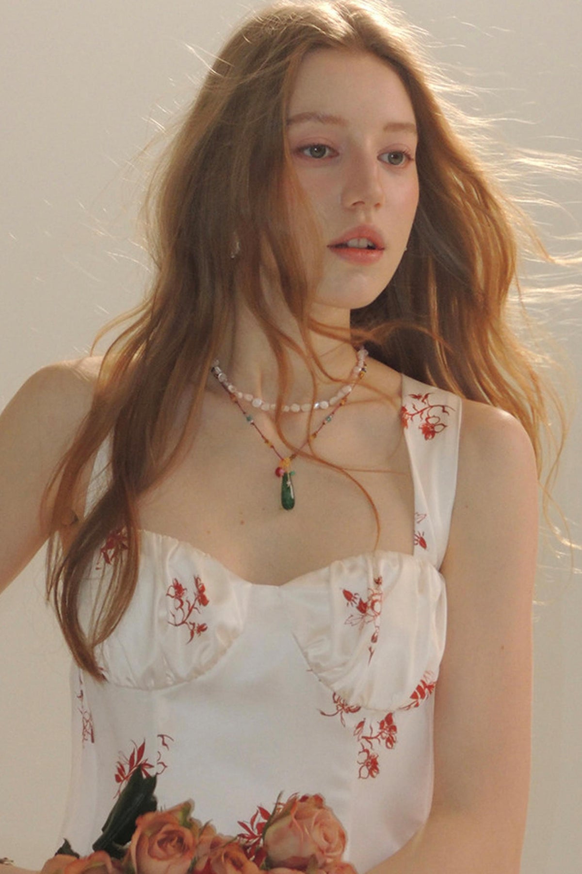 A woman with long wavy light brown hair wearing a white camisole top with red floral embroidery, layered necklaces, and holding a bouquet of pink roses against a warm off-white background.