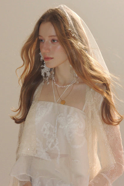 A woman wearing a white lace bridal-style top with a sheer veil and layered pearl necklaces, photographed against a soft warm beige background.