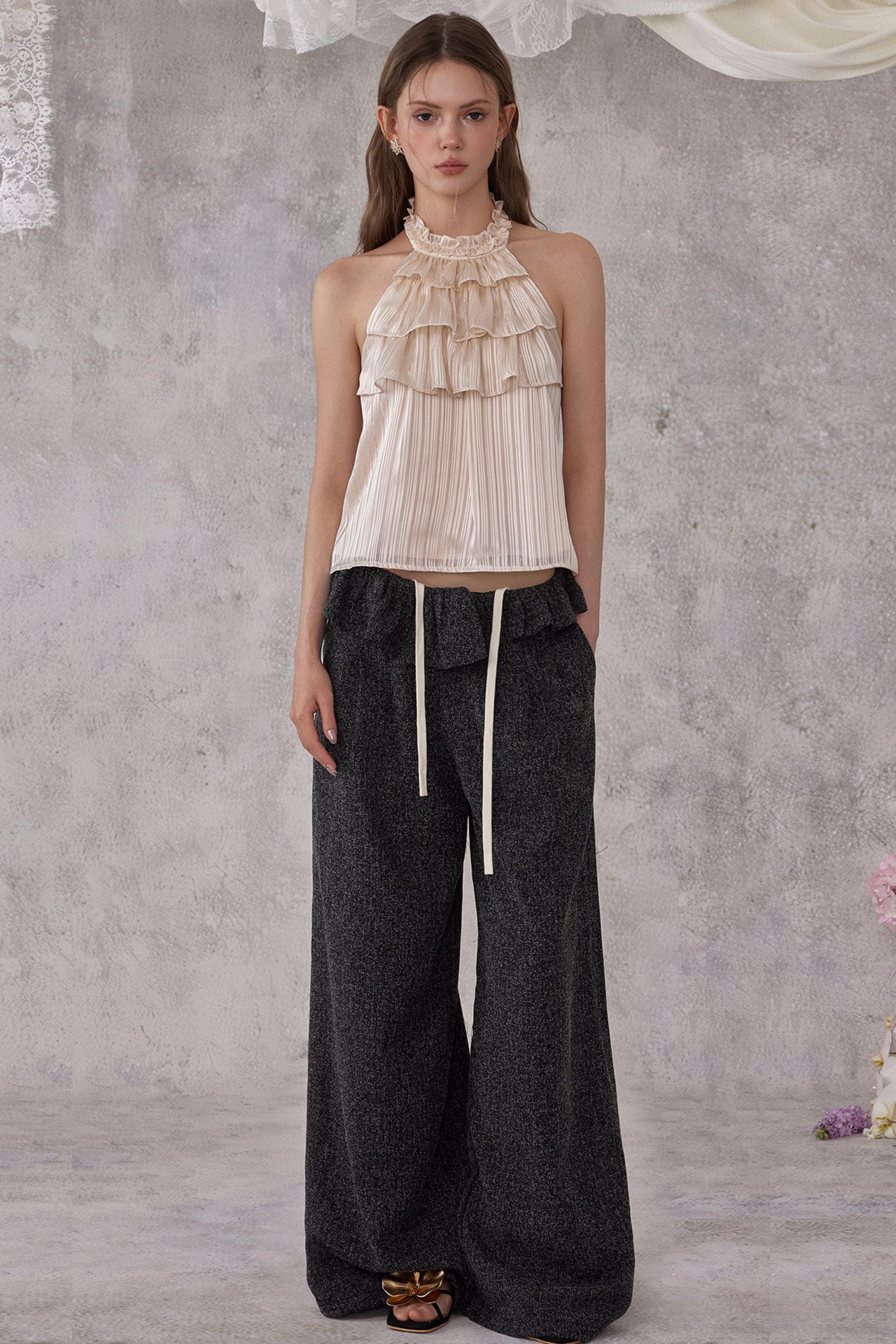 Woman wearing a beige ruffled top and dark wide-leg pants against a gray textured wall.