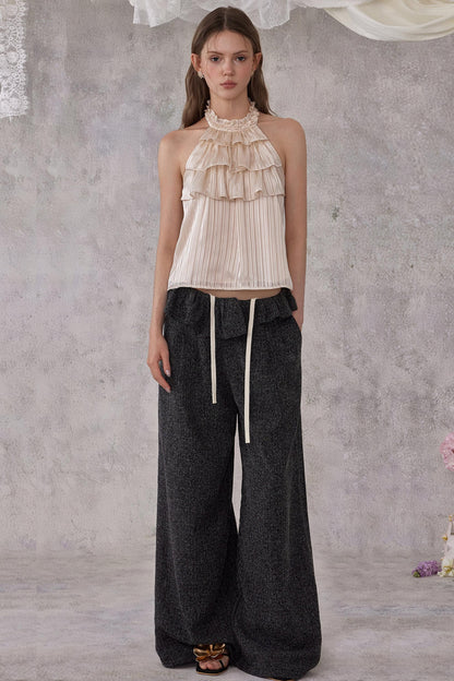 Woman wearing a beige ruffled top and dark wide-leg pants against a gray textured wall.