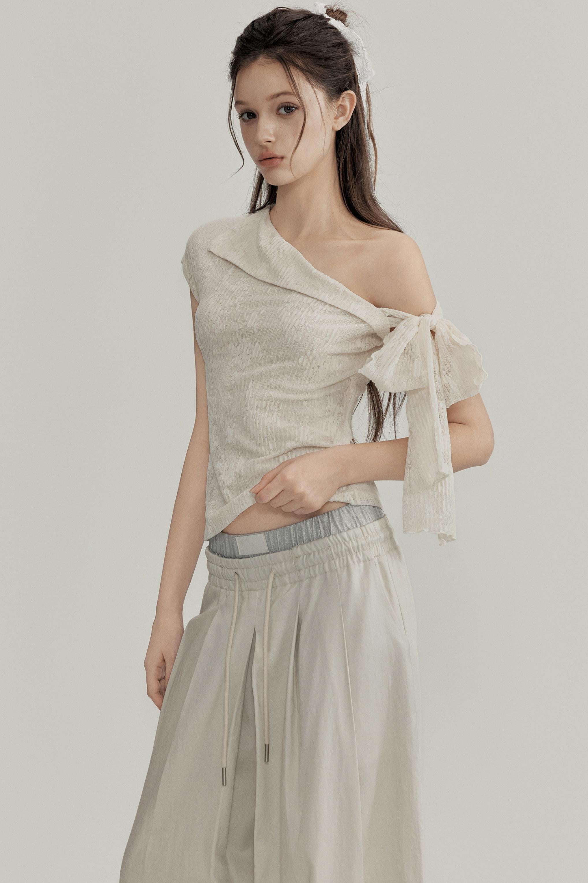 Woman wearing a light-colored one-shoulder top with a bow detail on a plain background