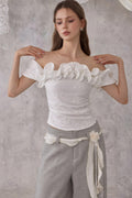 Woman wearing a white off-shoulder top with floral details and gray pants against a gray background