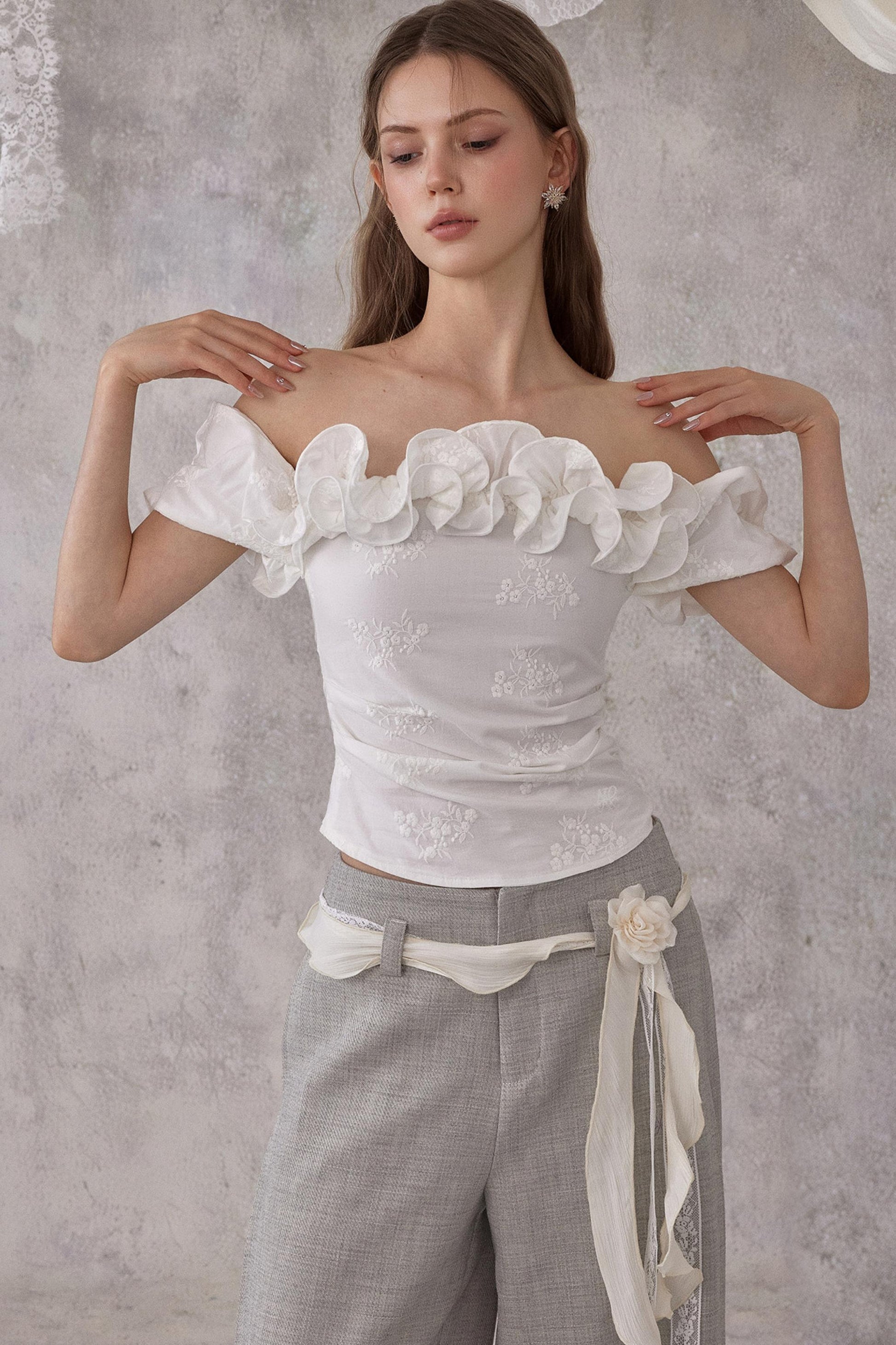 Woman wearing a white off-shoulder top with floral details and gray pants against a gray background