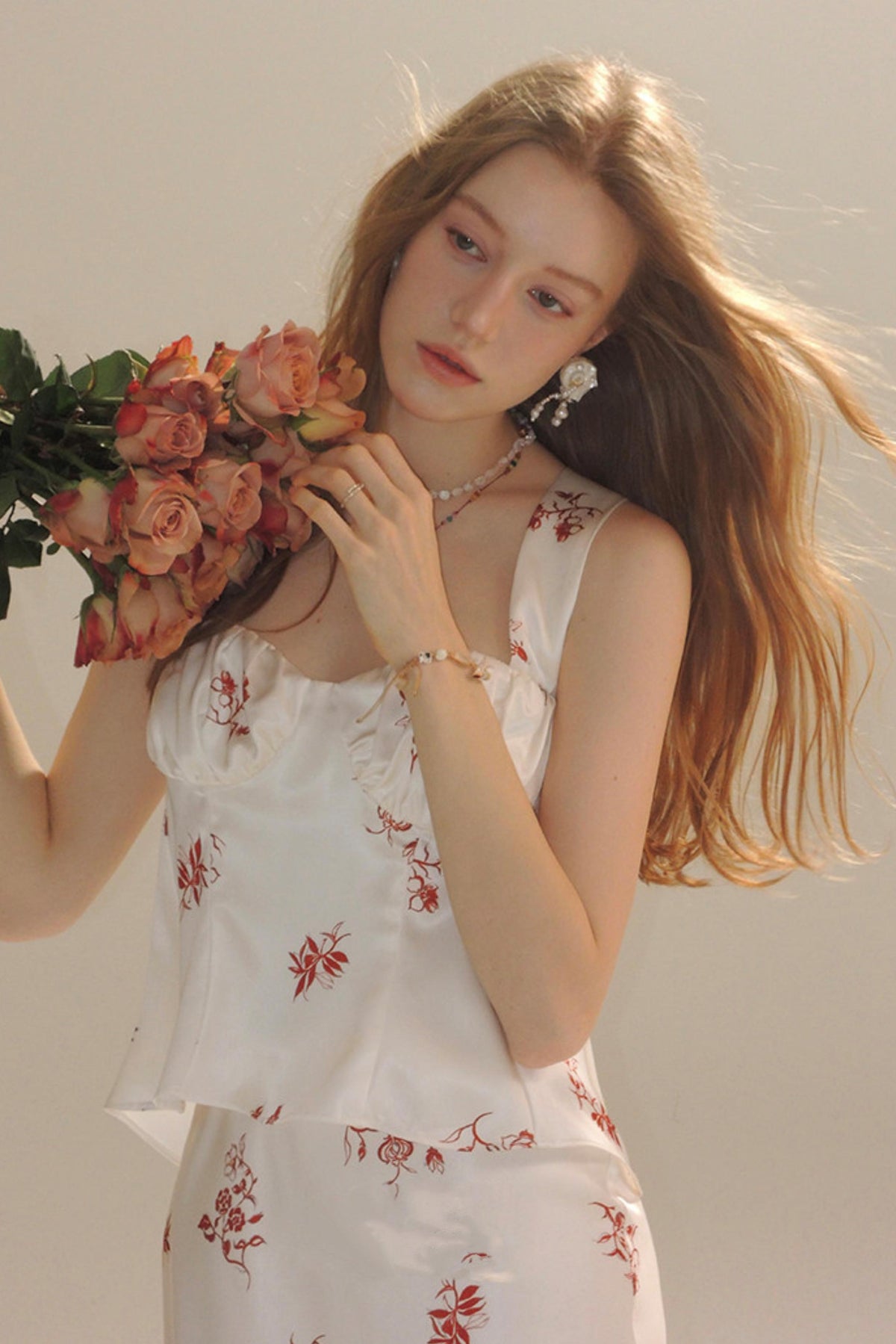 A woman with long light brown hair holds a bouquet of peach roses, wearing a white sleeveless top and matching bottoms with a red floral print, against a plain light beige background.
