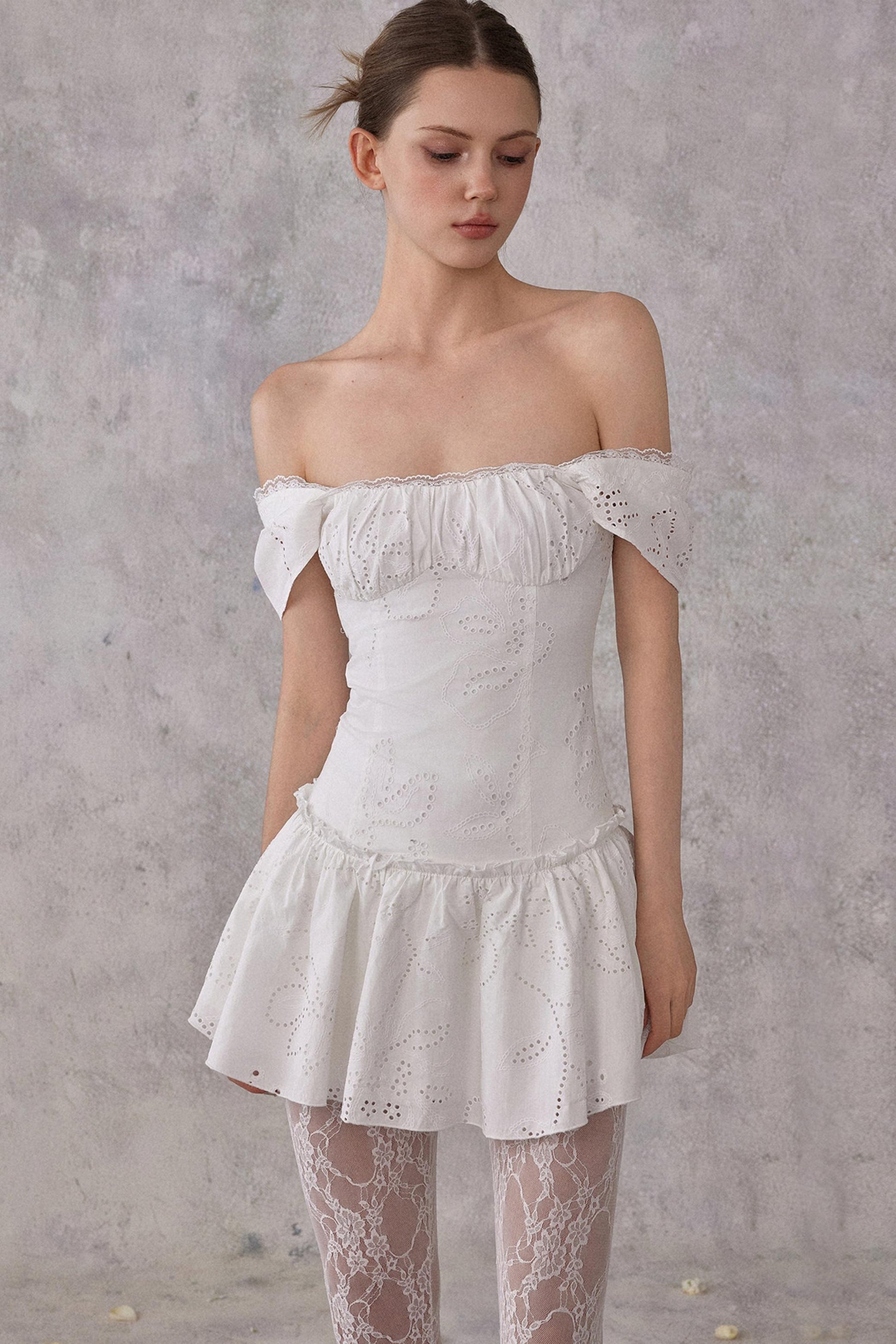 Model styling a romantic white cotton mini dress with eyelet lace details and a smocked pleated waist, worn in a feminine balletcore aesthetic.