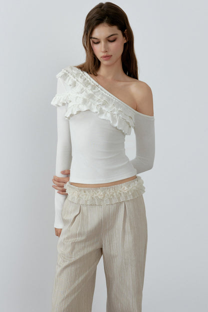Woman wearing a white one-shoulder top with ruffles and beige pants on a plain background