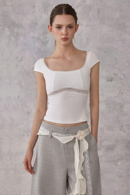 Woman wearing a white top with lace details and gray pants against a textured gray background