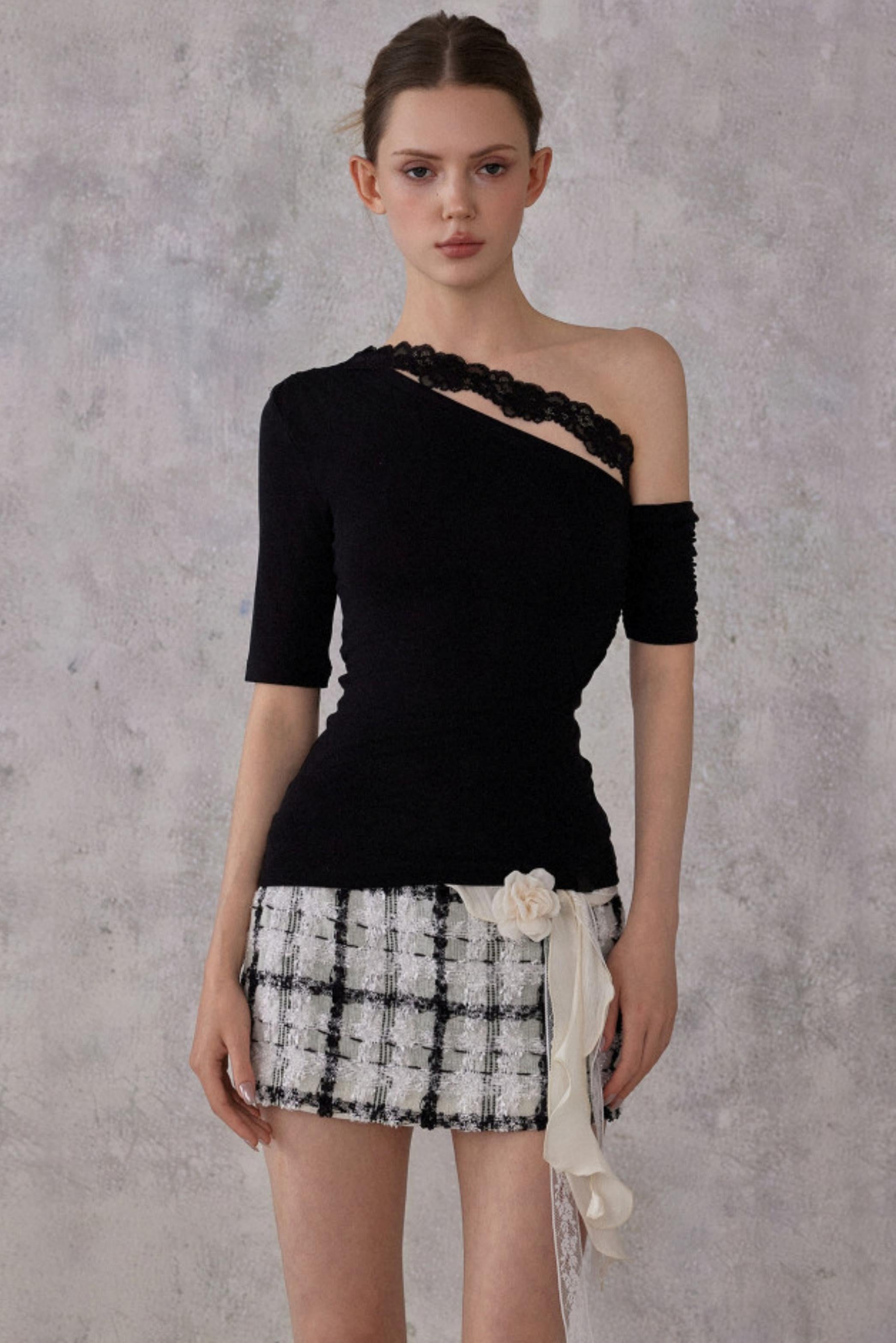 Model wearing a black one-shoulder top with a plaid skirt against a gray background