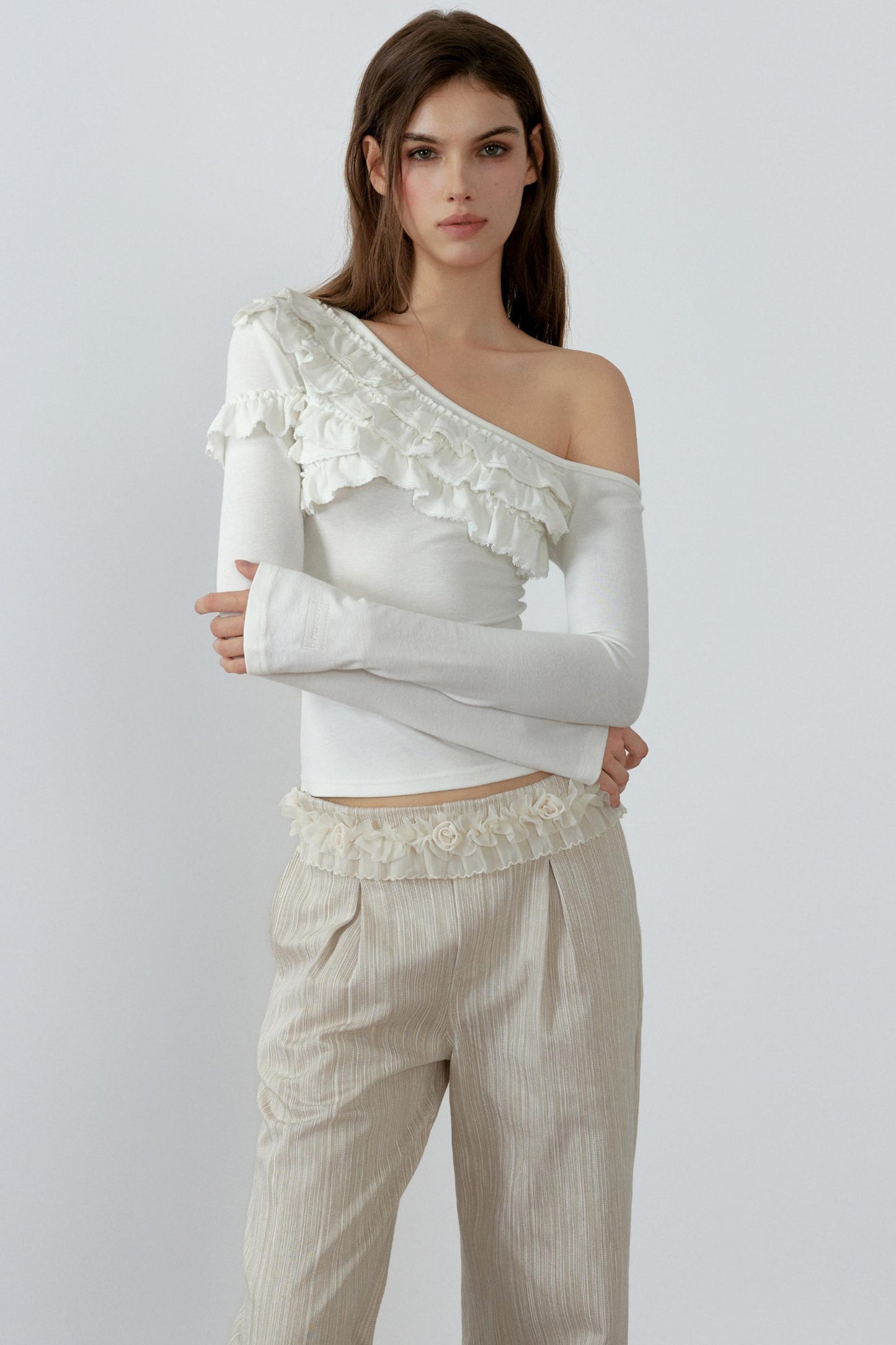 Woman wearing a white off-shoulder top with ruffled details and beige pants on a light gray background