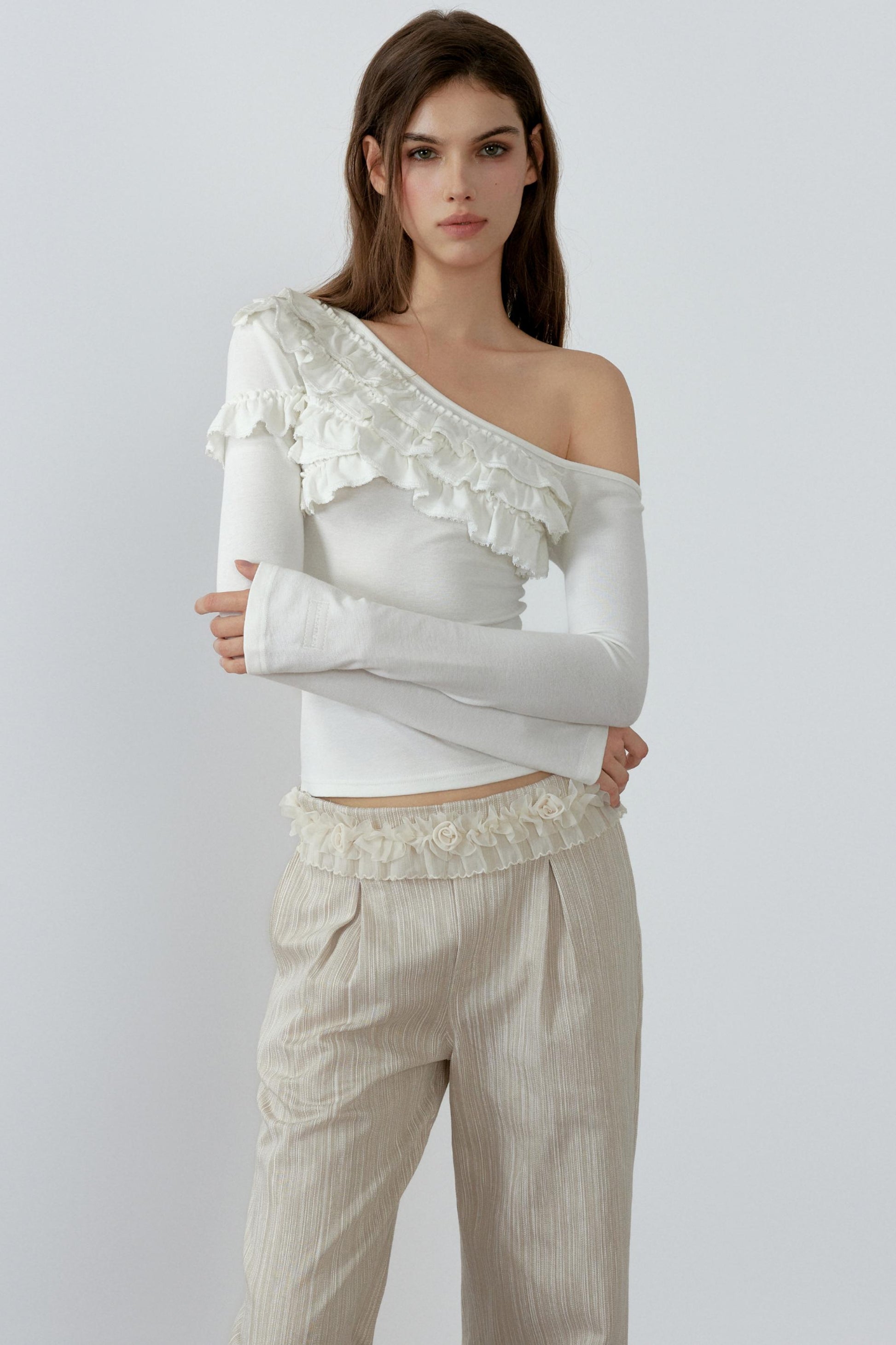 Woman wearing a white off-shoulder top with ruffled details and beige pants on a light gray background