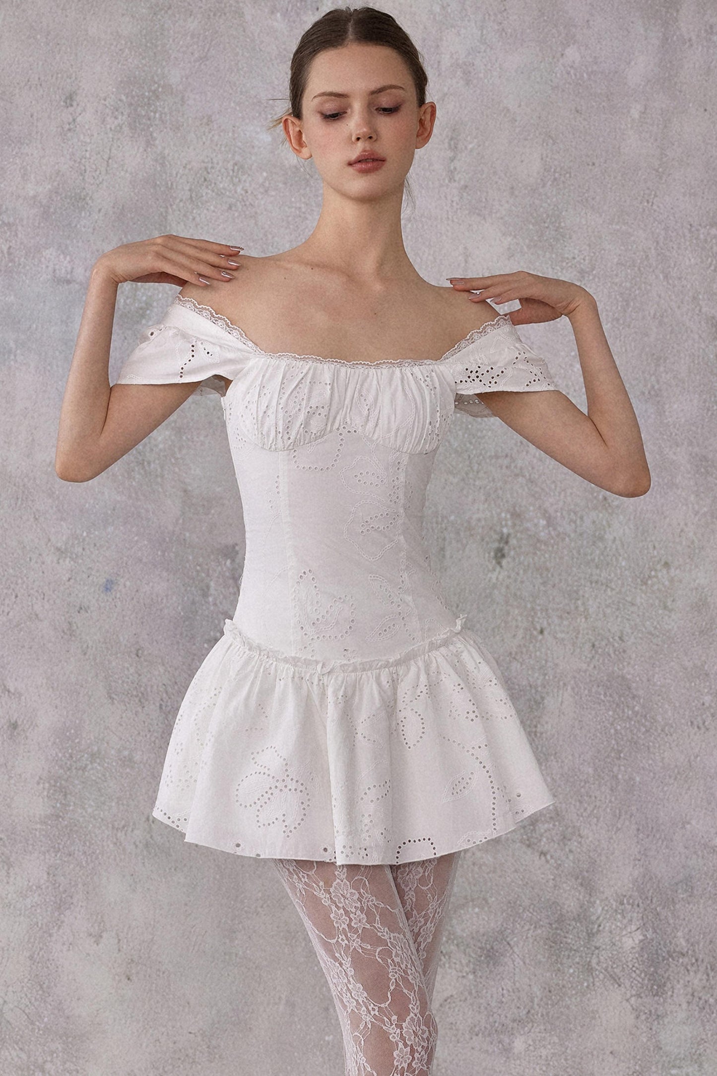 Model styling a romantic white cotton mini dress with eyelet lace details and a smocked pleated waist, worn in a feminine balletcore aesthetic.