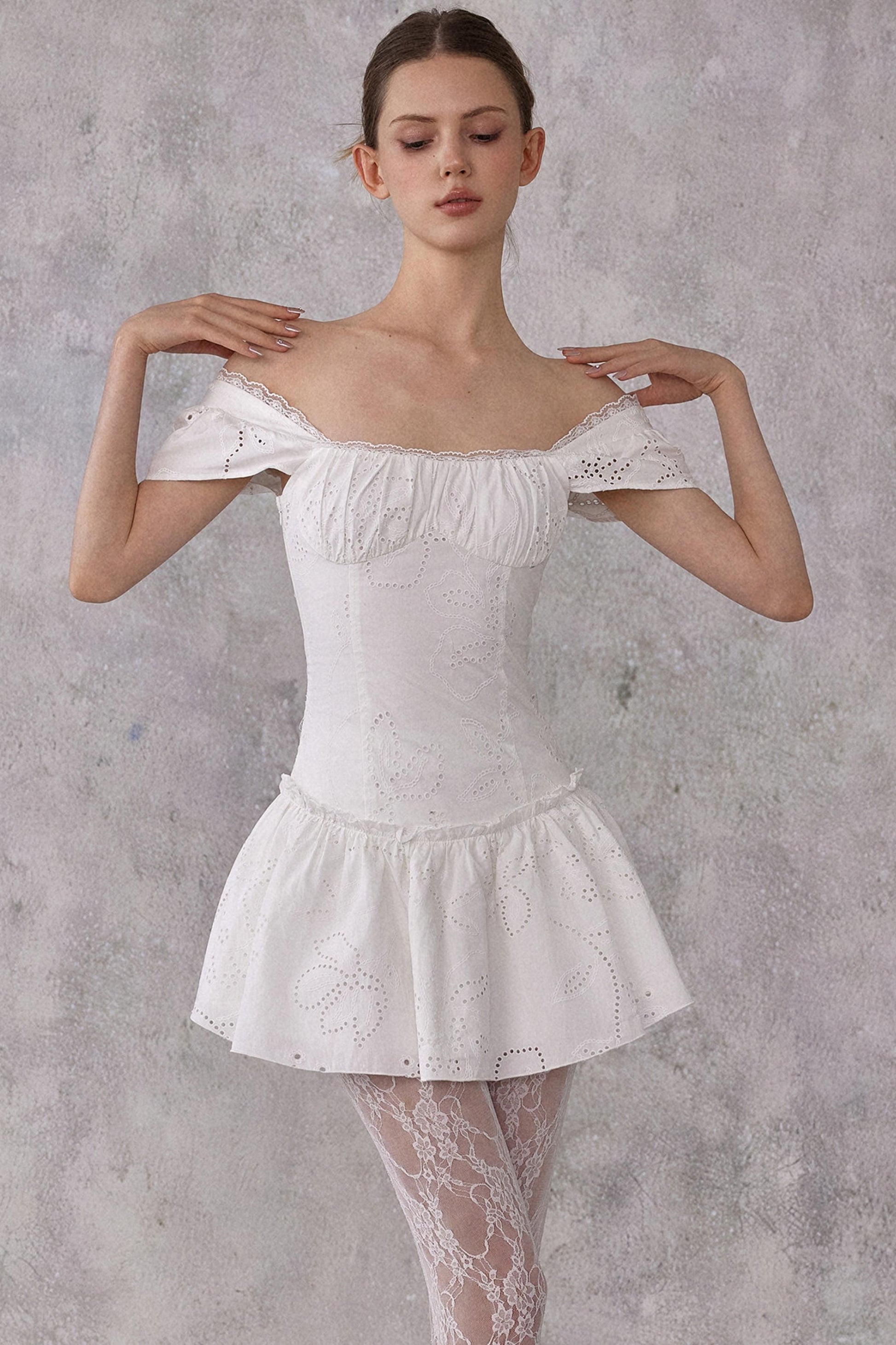 Model styling a romantic white cotton mini dress with eyelet lace details and a smocked pleated waist, worn in a feminine balletcore aesthetic.