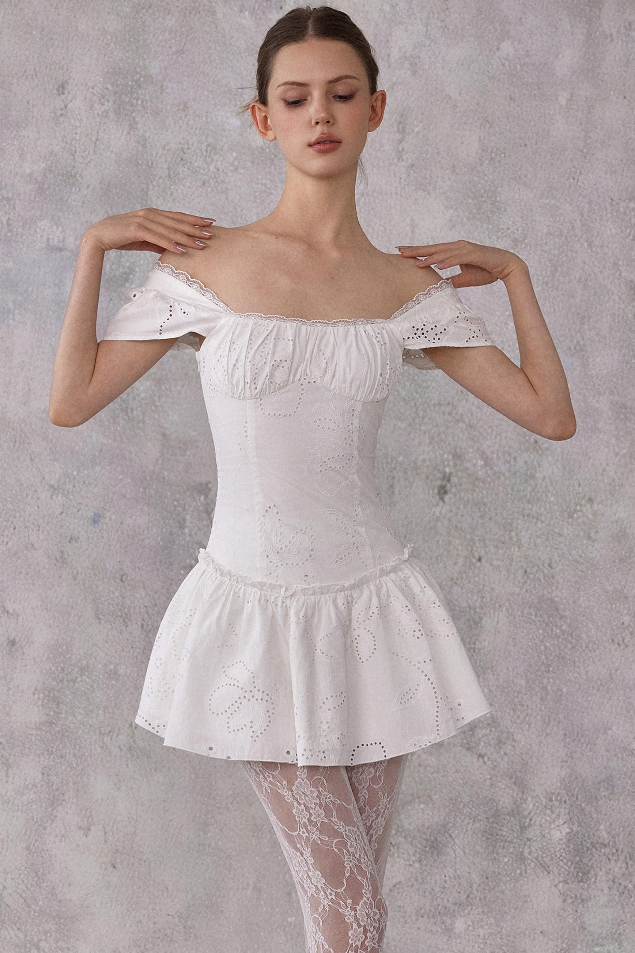 Model styling a romantic white cotton mini dress with eyelet lace details and a smocked pleated waist, worn in a feminine balletcore aesthetic.