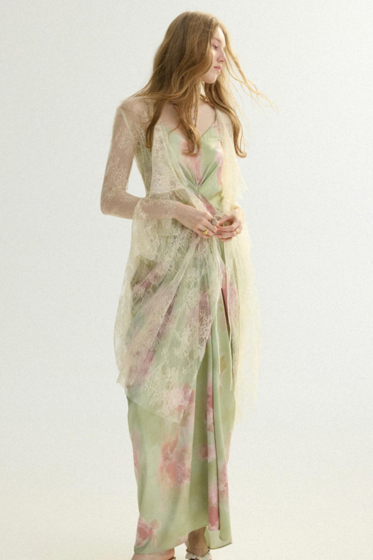 A woman modeling a long, sheer, light sage green maxi dress with a pink floral print and a matching draped lace overlay, set against a plain white studio background.