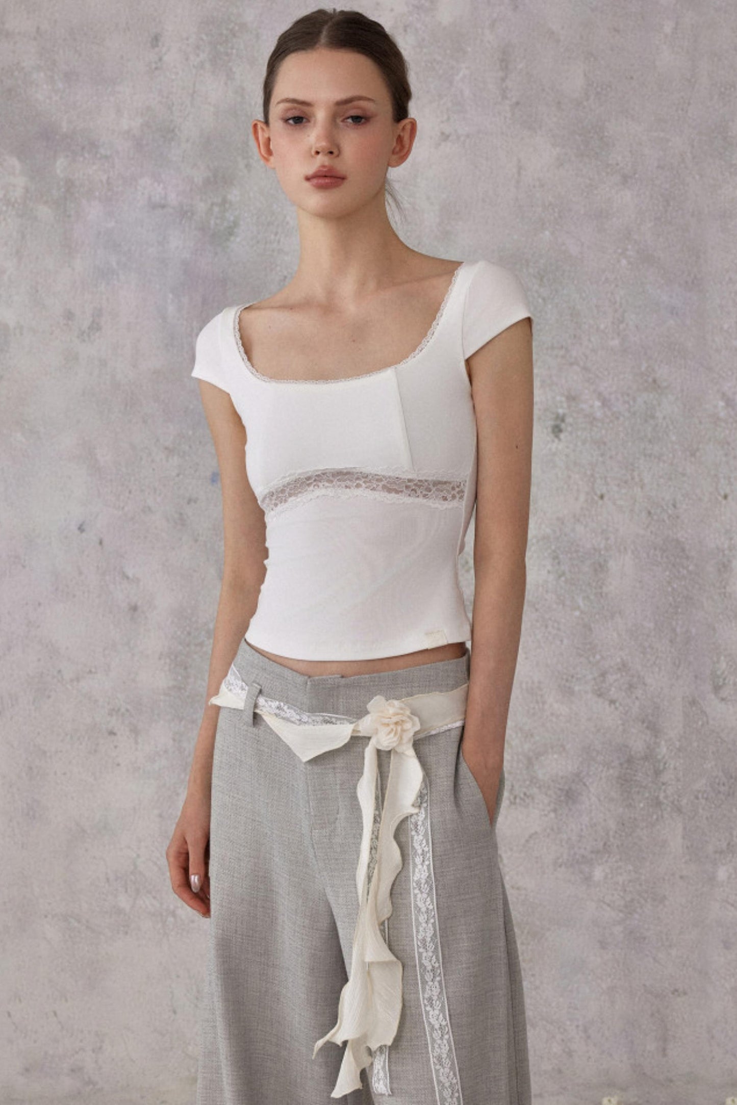 Woman wearing a white top and gray pants with lace details against a gray background