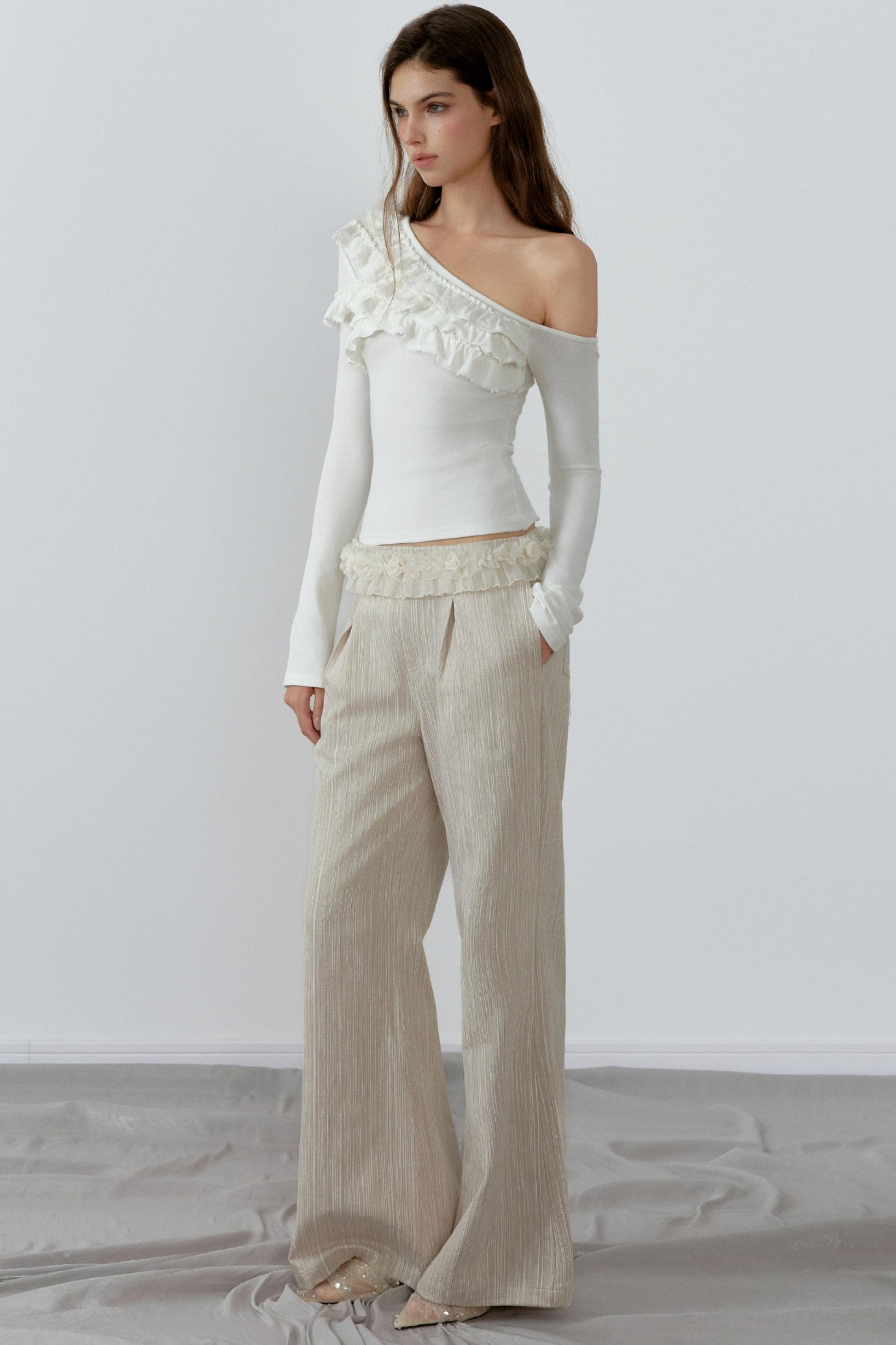 Woman wearing a white one-shoulder top and beige pants against a plain background