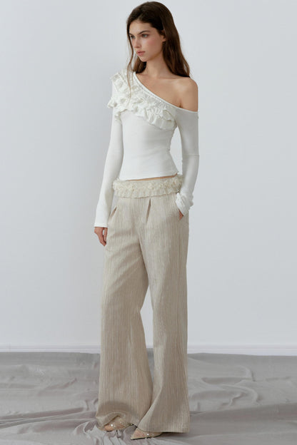 Woman wearing a white one-shoulder top and beige pants against a plain background