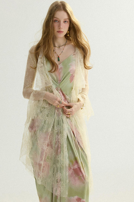 A woman modeling a light sage green satin dress with pink floral prints, layered under a sheer cream lace shawl, against a plain off-white background.