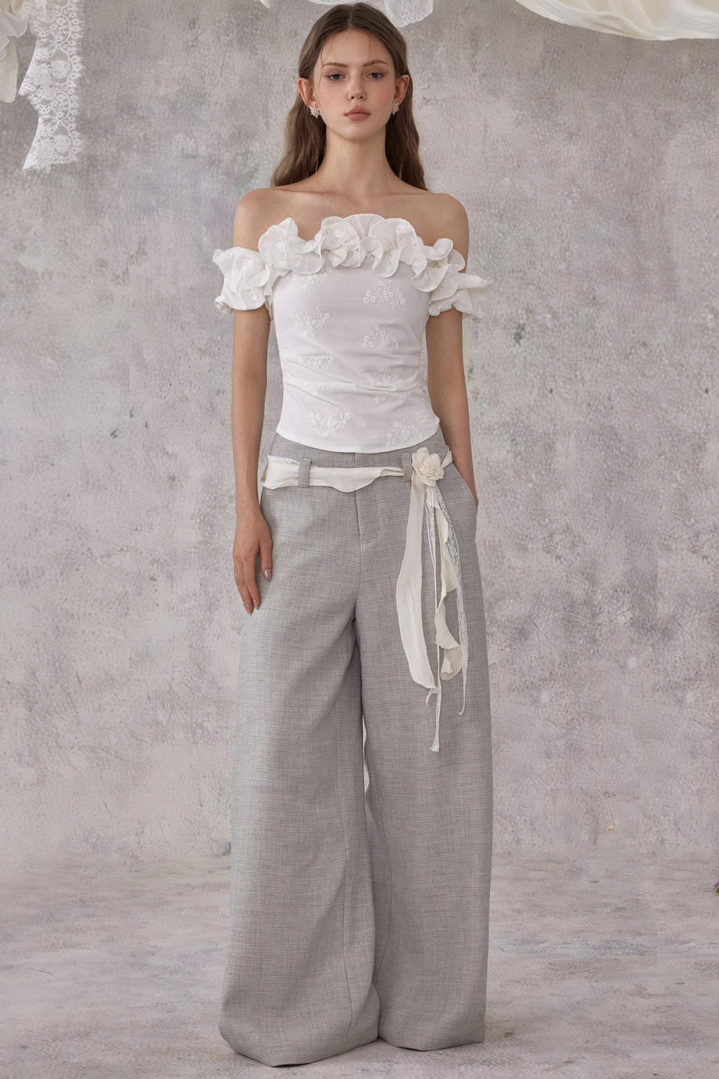 Woman wearing a white floral top and gray wide-leg pants against a textured gray background