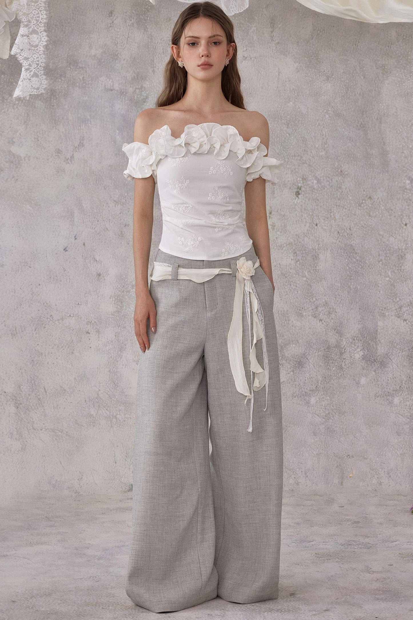 Woman wearing a white floral top and gray wide-leg pants against a textured gray background