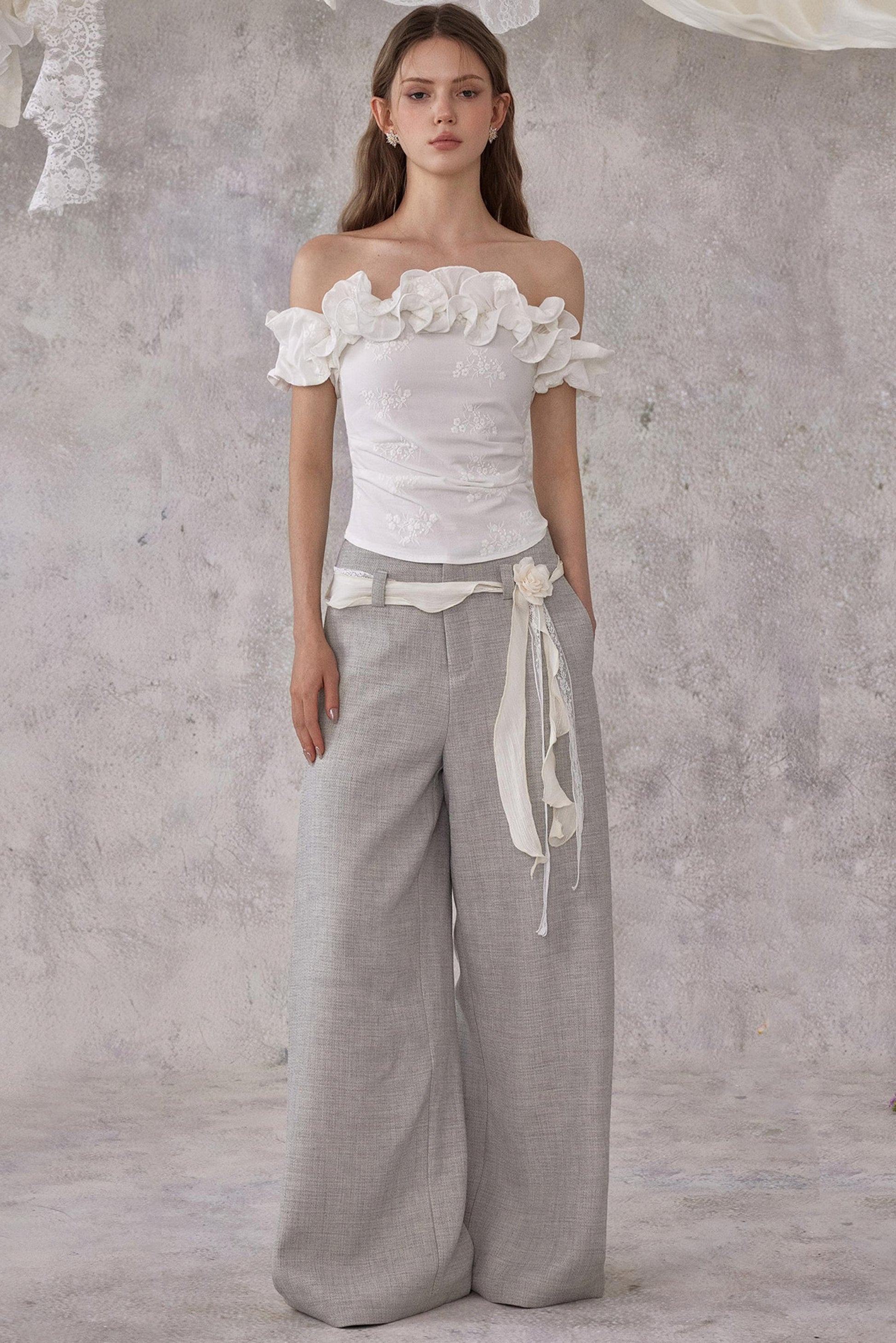 Woman wearing a white floral top and gray wide-leg pants against a textured gray background