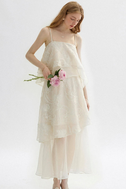 A woman wearing a cream-colored, tiered lace and tulle midi dress, holding a small bouquet of pink roses against a plain white studio background.