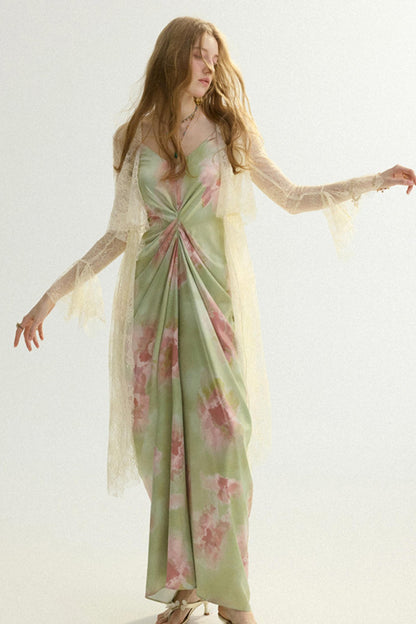 A woman modeling a long, flowing sage green dress with pink floral patterns and sheer cream lace sleeves, posed against a plain white studio background.