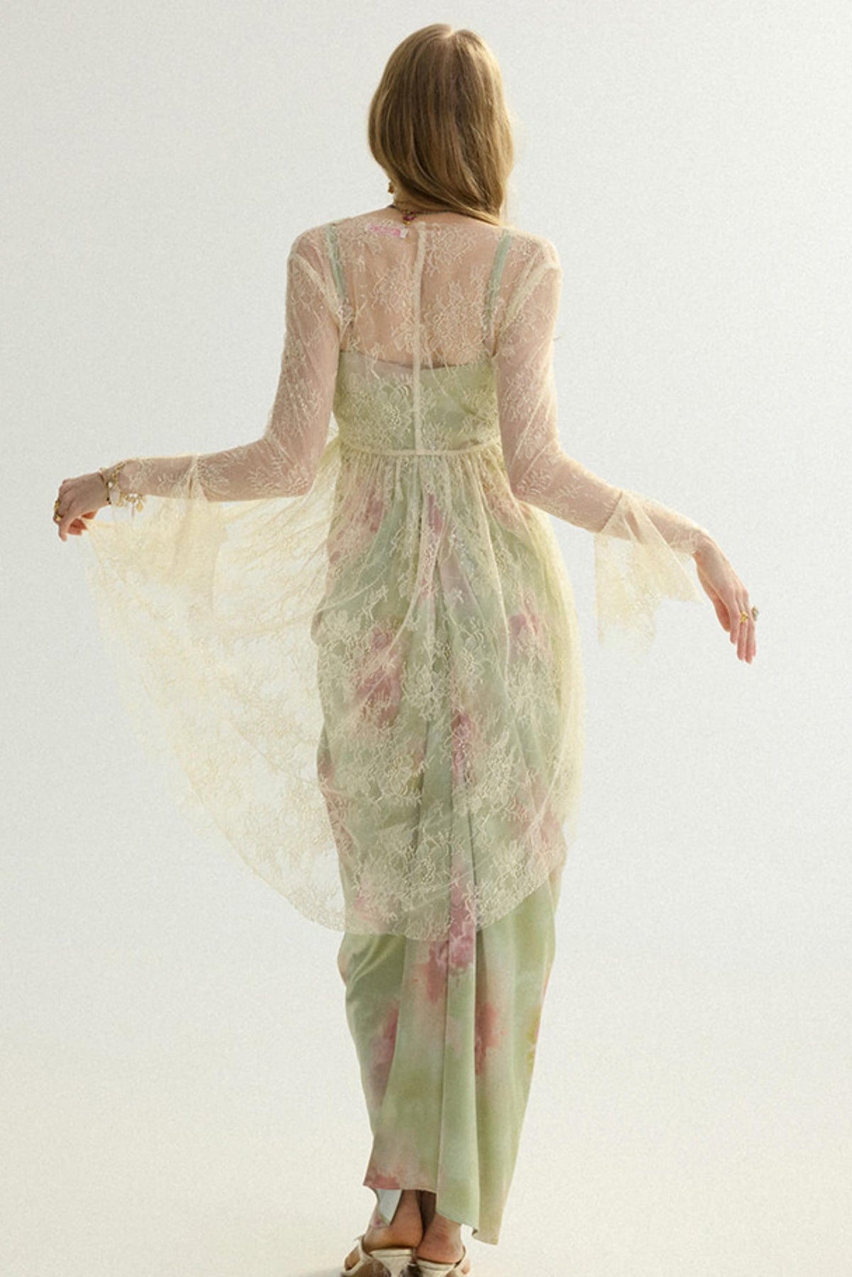 A woman viewed from behind wearing a flowing sheer long-sleeve dress in pale sage green with pink floral accents and a matching sheer overlay, photographed against a plain light background.