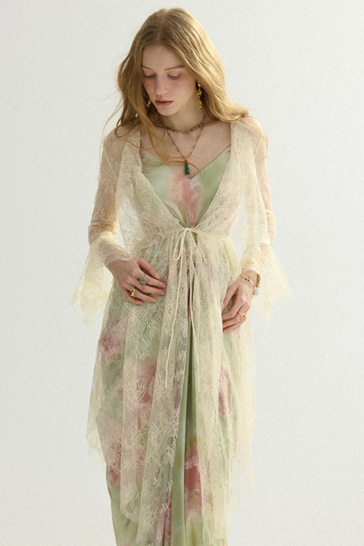 A woman wearing a sheer, long cream lace robe over a light sage green and pink floral dress, standing against a plain off-white background.