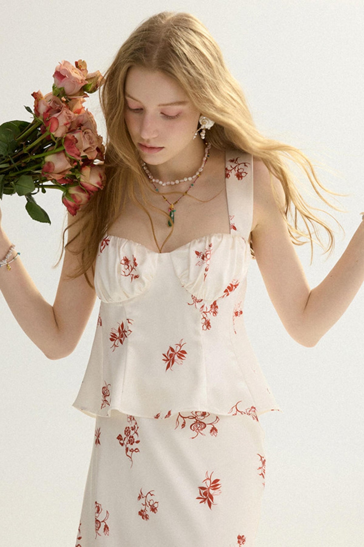 A blonde woman wearing a white sleeveless peplum dress with a red floral print, holding a bouquet of pink roses against a plain white background.