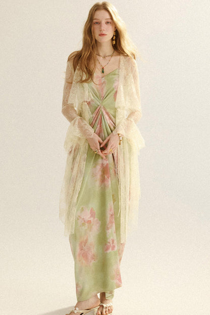 A woman modeling a light sage green maxi dress with pink floral patterns, layered under a sheer cream lace cardigan, against a plain light background.