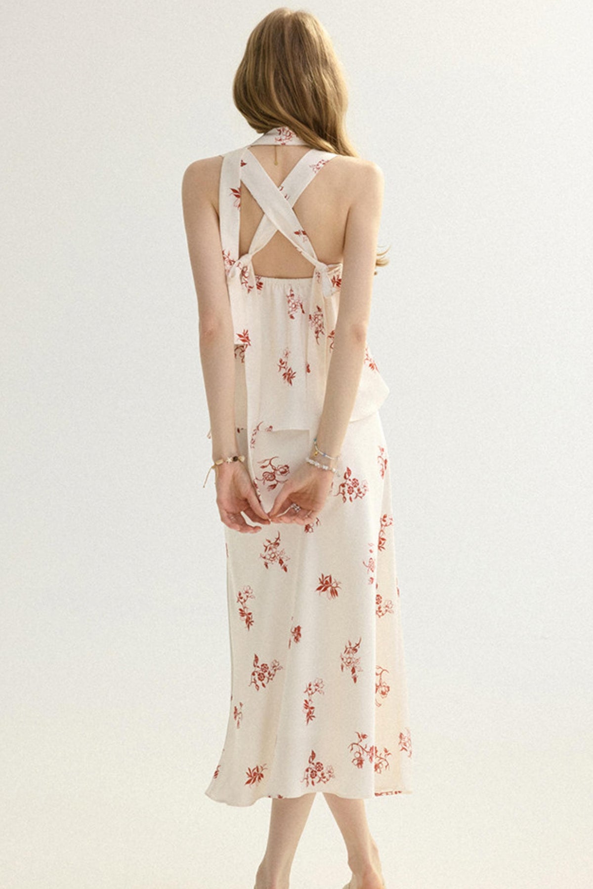 A woman shown from the back wearing a sleeveless cream dress with a red floral print and crisscross back straps, photographed against a plain light background.