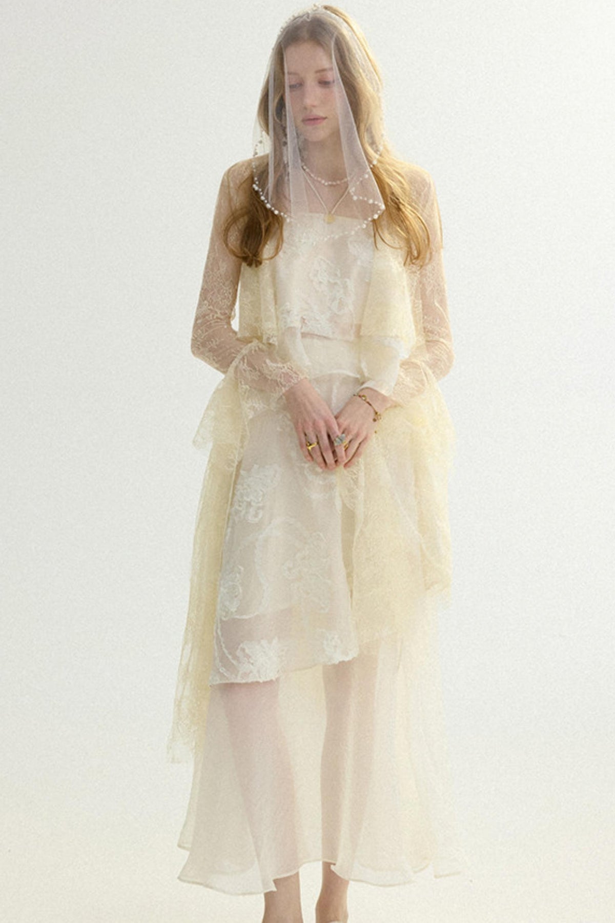 A woman wearing a long cream lace dress with sheer long sleeves, a matching veil, and a layered tulle overlay, photographed against a plain light background.