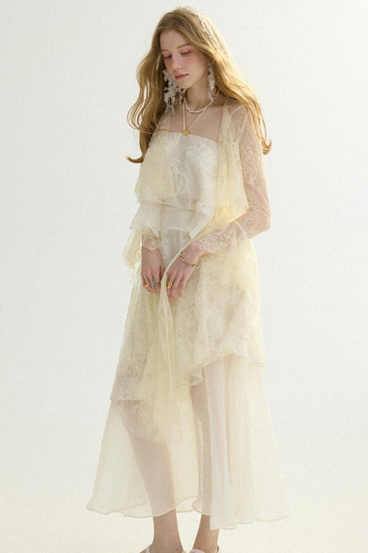 A woman modeling a long cream-colored dress with a sheer lace overlay and ruffled layers, photographed against a plain light background.