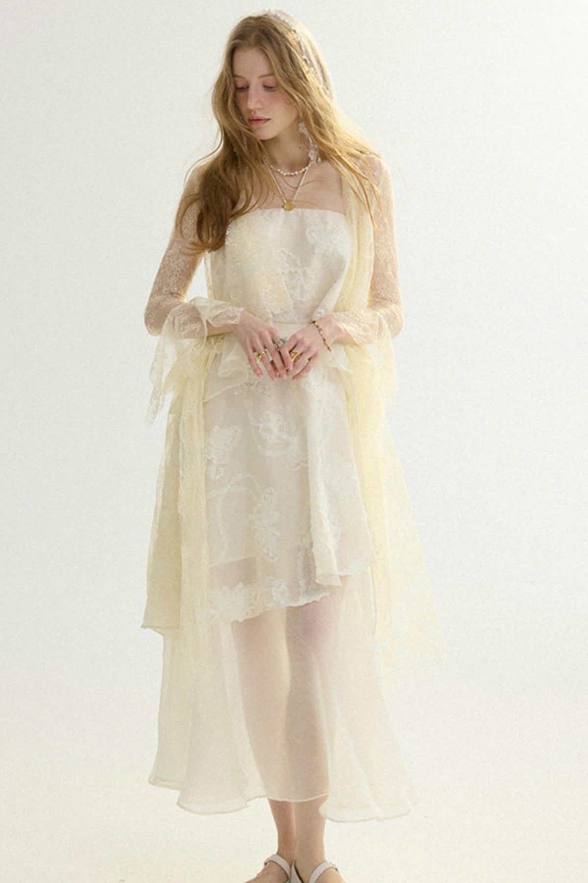 A woman modeling a cream-colored sheer lace dress with a matching long-sleeve tulle shawl, set against a plain light background.