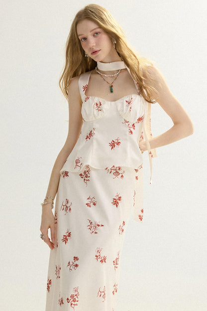 A woman modeling a sleeveless white midi dress with a red floral print, featuring a ruffled bust and tie straps, set against a plain white studio background.