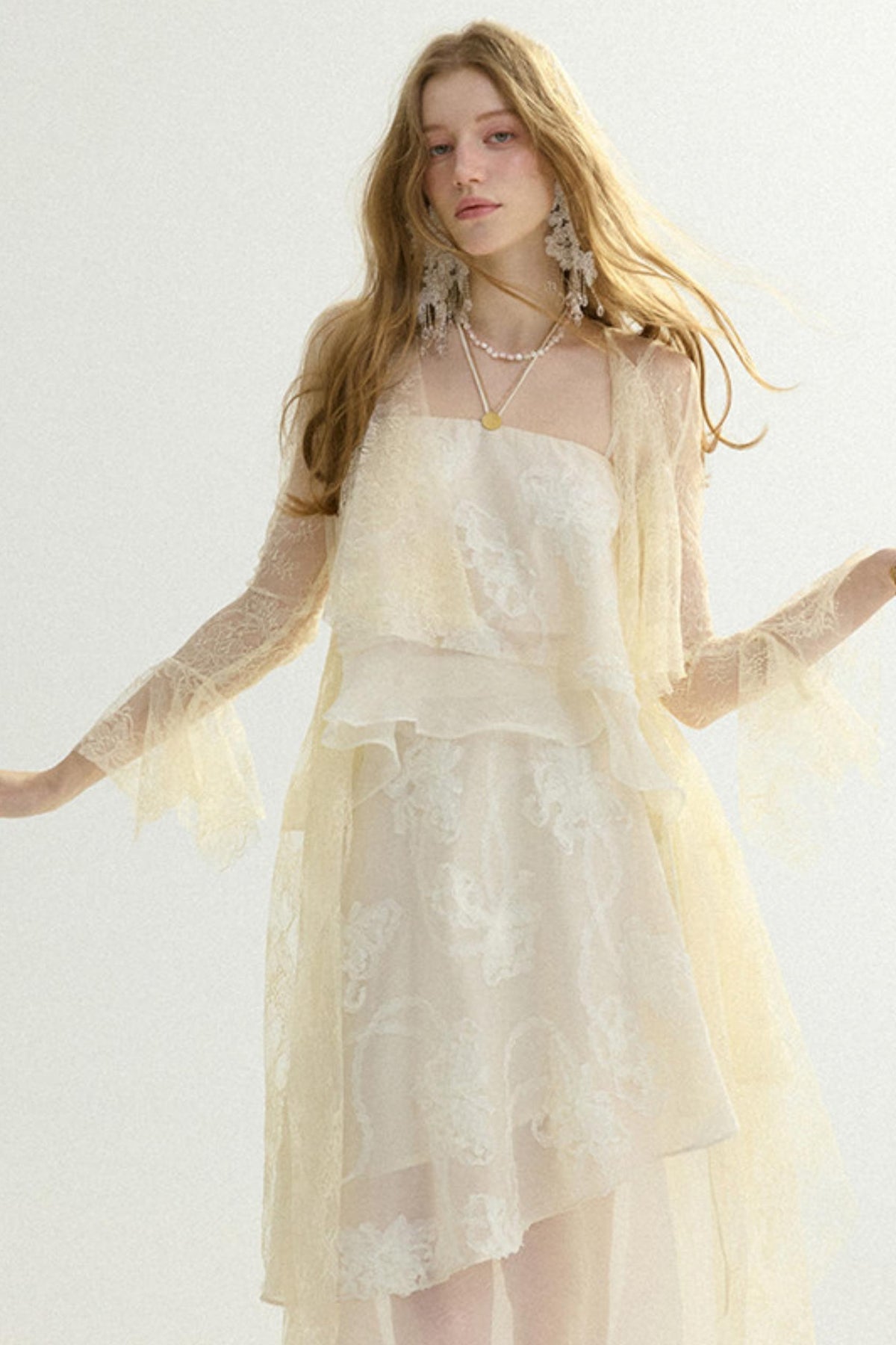 A woman modeling a cream-colored lace dress with sheer long sleeves and layered ruffles, set against a plain light background.