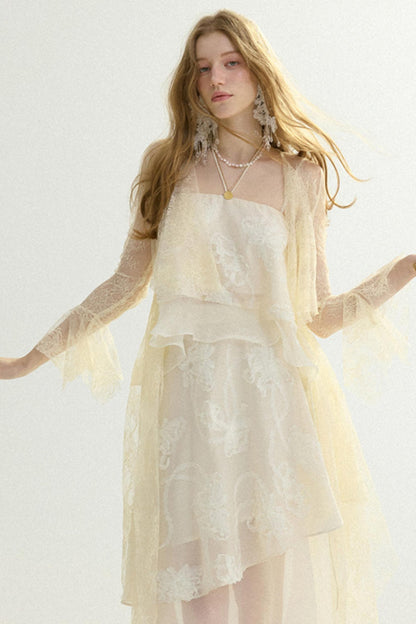 A woman modeling a cream-colored lace dress with sheer long sleeves and layered ruffles, set against a plain light background.